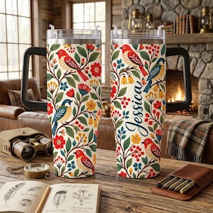 May include: Two stainless steel tumblers with handles, featuring a cream background with a colorful pattern of red and blue birds, green leaves, and red and yellow flowers. The right tumbler displays the name Jessica in a dark blue script font.