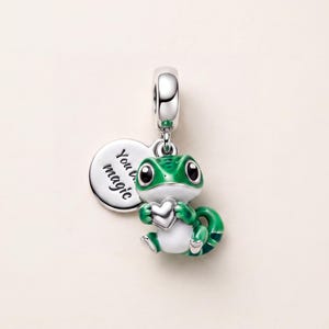 May include: A silver charm featuring a green and white lizard holding a small silver heart. Attached to the charm is a round silver disc engraved with the text You are magic. The lizard has large black eyes and a detailed green textured back.