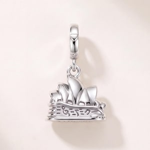 May include: A silver charm shaped like the Sydney Opera House, featuring the iconic sail-like roof structures and a base engraved with musical notes and the text "SYDNEY". The charm includes a circular bail at the top for attachment to a bracelet.