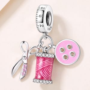 May include: A silver charm featuring a measuring tape bail marked with numbers 50, 60, 70, 80 and S925. Three dangling charms hang from the bail: a pair of silver scissors with three pink stones, a pink thread spool with clear stones, and a pink button.