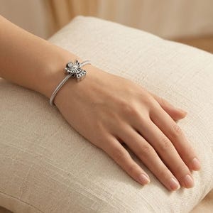 以下が含まれることがあります： 中央に天使の形のチャームが付いたシルバーカラーのチャームブレスレット。天使のチャームは、クリアストーンのアクセントが付いた円形の頭部と外側に広がる翼が特徴で、柔軟な質感のヘビ革風チェーンブレスレットに取り付けられています。
