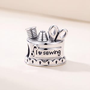 May include: A silver-toned metal charm featuring a sewing theme. The top displays a spool of thread, a thimble, and a pair of scissors. The front side includes the text "I heart sewing" with a heart symbol replacing the word heart.