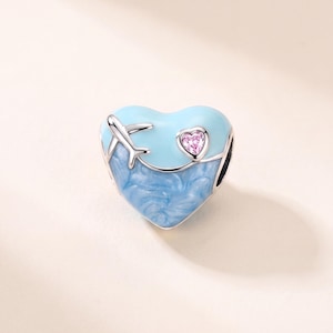 May include: A heart-shaped charm bead featuring a smooth light blue upper section with a silver aeroplane silhouette and a small pink heart-shaped crystal, and a textured, swirling blue lower section, all set within a polished silver metal frame.