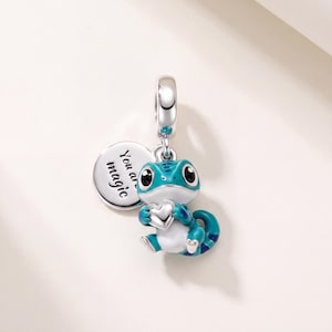 Può includere: Un ciondolo in argento a forma di lucertola blu e bianca che tiene un piccolo cuore. Al ciondolo è attaccato un disco rotondo in argento con inciso il testo 'You are magic'. La lucertola ha grandi occhi neri e una coda arricciata con una maglia.