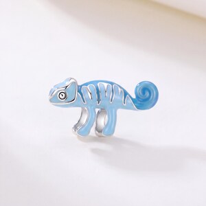 Puede incluir: Un dije de metal plateado con forma de camaleón, que presenta detalles de esmalte azul claro en el cuerpo. El diseño incluye una cola enroscada, cuatro patas y un ojo distintivo, con contornos plateados que separan las secciones de esmalte.
