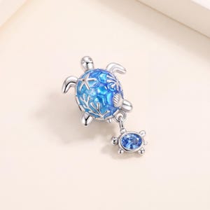 May include: A silver charm featuring a sea turtle with a blue enamel shell decorated with raised starfish and coral motifs. A smaller, dangling silver charm shaped like a baby turtle with a single blue crystal centre is attached to the main piece.