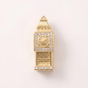 May include: A gold-toned charm shaped like the Elizabeth Tower, commonly known as Big Ben. The clock face is surrounded by a square border of small, clear, sparkling stones, with additional stones lining the base of the tower structure.