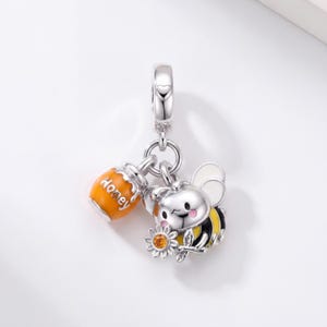 Puede incluir: Un dije de plata con una abeja de dibujos animados con rayas amarillas y negras, alas blancas y mejillas rosadas, que sostiene una pequeña flor con un centro naranja. Junto a la abeja hay un pequeño tarro de miel naranja con la palabra Honey.