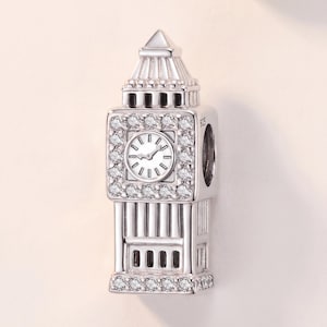 May include: A silver charm shaped like the Big Ben clock tower, featuring a central clock face surrounded by clear, sparkling stones. The piece is crafted with detailed architectural columns and a pointed roof, with the hallmark 925 visible on the side.