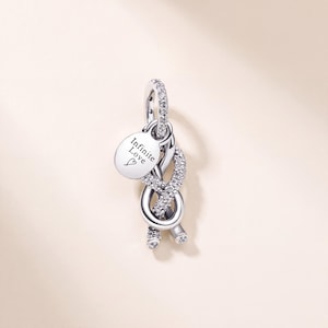 May include: A silver-toned charm featuring a knotted design encrusted with clear stones. A small, round, flat silver-toned tag hangs from the top loop, engraved with the text "Infinite Love" and a small heart symbol below the lettering.
