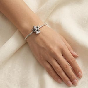 以下が含まれることがあります： 中央に天使の形のチャームが付いたシルバーカラーのスネークチェーンブレスレット。チャームには切り抜かれた翼の模様が施され、天使の体の中心には小さな透明な丸い宝石がセットされており、磨き上げられたメタリックな外観を演出しています。