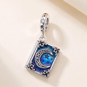 Puede incluir: Un charm de plata con forma de libro rectangular y cubierta de esmalte azul con un diseño de luna creciente y estrella. El charm incluye pequeñas piedras negras en las esquinas y una anilla decorada con estrellas grabadas y una piedra clara. S925 es visible.