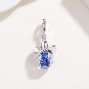 May include: A silver charm shaped like a pointed hat with a wide brim, featuring a spherical blue and white swirled glass bead at the base. The metal hat is accented with small embedded orange and blue gemstones along the top and the bail.