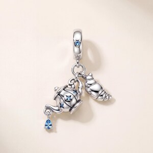 May include: A silver charm featuring a teapot and a croissant dangling from a bail. The teapot is decorated with small blue star-shaped stones and has a teardrop-shaped blue stone hanging from the spout. The bail also features a small blue heart.