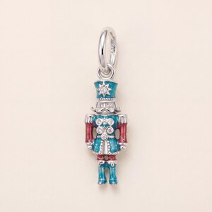 May include: A sterling silver nutcracker charm featuring teal and red enamel detailing with a glittery finish. The figure is adorned with small clear stones set into the hat, face, and jacket buttons. The piece includes a circular jump ring.