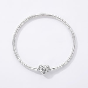 May include: A silver snake chain bracelet featuring a heart-shaped clasp. The clasp is decorated with an intricate, engraved floral pattern and small embedded stones. The bracelet has a smooth, flexible, and rounded metallic texture throughout.