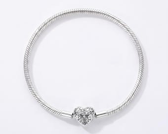 May include: A silver snake chain bracelet featuring a heart-shaped clasp. The clasp is decorated with an intricate, engraved floral pattern and small embedded stones. The bracelet has a smooth, flexible, and rounded metallic texture throughout.