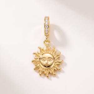 Peut inclure: Un pendentif doré représentant un visage de soleil central avec des rayons ondulés rayonnants. La bélière circulaire au sommet est ornée d'une rangée verticale de petites pierres claires et scintillantes, contrastant avec le métal poli.