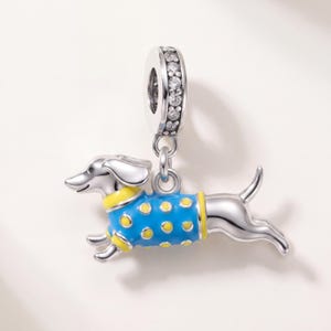 May include: A silver charm shaped like a dachshund wearing a blue jumper with yellow polka dots and yellow trim. The charm features a cylindrical bail encrusted with small clear stones, and the dog is depicted in a running pose.