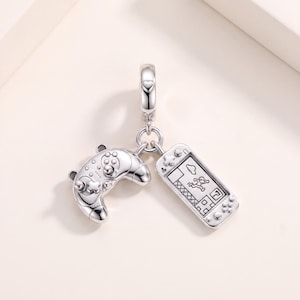 May include: A silver charm featuring a handheld gaming console and a game controller suspended from a small ring. The console displays a screen with a character running through a platform game level. The metal has a polished, reflective finish.