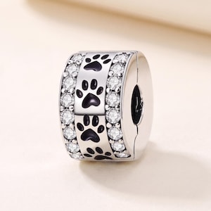 May include: A silver-toned charm bead featuring a central band decorated with black paw print motifs, flanked on both sides by rows of clear, round-cut sparkling stones. The cylindrical piece has a polished metallic finish and a hollow center.