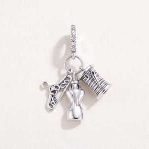 May include: A silver charm featuring a circular bail with embedded stones, suspending three distinct pendants: a filigree clothes hanger with a small stone, a smooth dress form mannequin, and a spool of thread with a needle resting on its side.