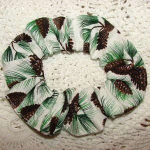 May include: A white scrunchie with a repeating pattern of brown pine cones and green pine needles. The fabric has a subtle silver shimmer. This hair accessory is perfect for adding a touch of nature-inspired style.