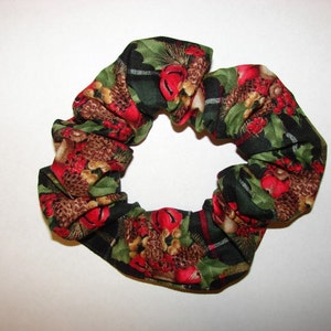 May include: A fabric scrunchie with a festive holiday pattern. The scrunchie features a repeating design of red berries, pinecones, and green holly leaves on a black background with plaid accents. The elastic hair accessory is circular.