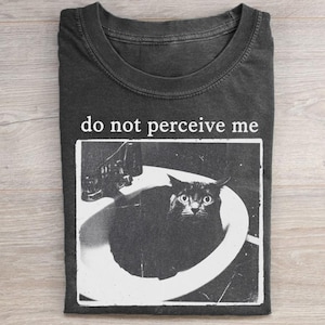 May include: A charcoal gray crew neck t-shirt featuring a white graphic print of a black cat sitting inside a bathroom sink. Above the graphic, the text "do not perceive me" is printed in a white, serif font. The shirt has a short-sleeved design.