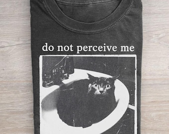 Peut inclure: Un t-shirt gris anthracite à col rond avec une impression graphique blanche représentant un chat noir assis dans un lavabo. Au-dessus du graphique, le texte "do not perceive me" est imprimé dans une police serif blanche. T-shirt à manches courtes.