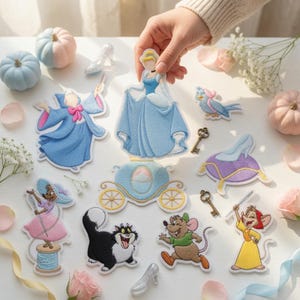 May include: A collection of embroidered fabric patches featuring characters and objects from the Cinderella story, including a blue-gowned princess, a fairy godmother, a pumpkin carriage, mice, a cat, a glass slipper on a pillow, and two keys.