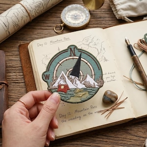 May include: Embroidered patch featuring a compass design with a mountain scene and the text "Day 12: Mountain Pass". The patch is in shades of green, white, and red. The patch is held over an open journal with a map and pen.
