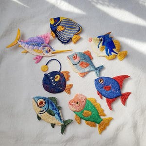 May include: A collection of colorful, embroidered fish patches in various shapes and sizes. The patches feature detailed stitching and vibrant colors, including blue, yellow, pink, and green. The fish designs include a swordfish, a blue tang, and an anglerfish.