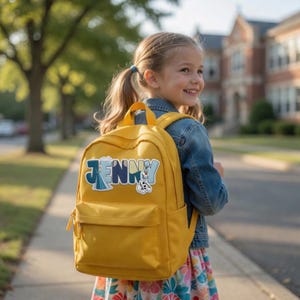 Puede incluir: Una mochila de color amarillo mostaza con un bolsillo frontal y un parche personalizado con el nombre JENNY en letras azules, decorada con gr&aacute;ficos de dibujos animados de una reina y un mu&ntilde;eco de nieve integrados en las letras del panel frontal.