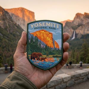 May include: A shield-shaped patch featuring the text "YOSEMITE NATIONAL PARK" above a landscape scene. The scene depicts a mountain with a waterfall, trees, and two bears in a river. The patch has a green border and is held in a hand.