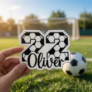 Puede incluir: Un parche bordado en blanco y negro con forma del número 22 y un patrón de balón de fútbol. El nombre Oliver está bordado en letra cursiva negra en la parte inferior del parche, que cuenta con un borde blanco y un reverso blanco.