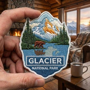May include: An embroidered patch featuring a mountain landscape with a brown bear walking near a blue lake, surrounded by green pine trees. The text GLACIER NATIONAL PARK is printed in white on a blue rectangular background at the bottom.