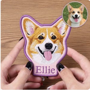 Könnte beinhalten: Gestickter Aufnäher mit einem lächelnden Corgi-Gesicht mit rosa Zunge. Der Aufnäher hat einen lila Rand und den Namen "Ellie" in Lila gestickt. Ein kleineres rundes Bild des Hundes befindet sich in der oberen rechten Ecke.