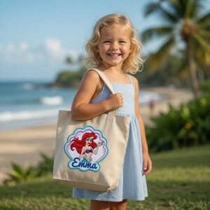 May include: A beige canvas tote bag featuring a colorful embroidered graphic of a red-haired mermaid inside a scalloped shell shape. The name Emma is embroidered in blue script lettering below the mermaid figure on the front of the bag.