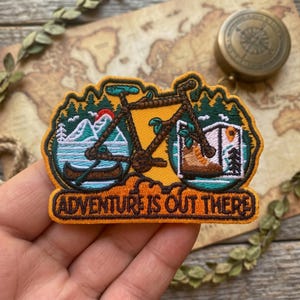 May include: Embroidered patch with a yellow background and a brown bicycle design. The patch features a canoe, mountains, a hiking boot, and the text "ADVENTURE IS OUT THERE". The patch is surrounded by a green border.