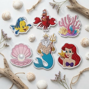 May include: A collection of embroidered patches featuring a yellow fish, a red crab, pink coral, a pink seashell with a white pearl, a merman with a gold trident, and a red-haired character holding a yellow starfish, all arranged on a white surface.