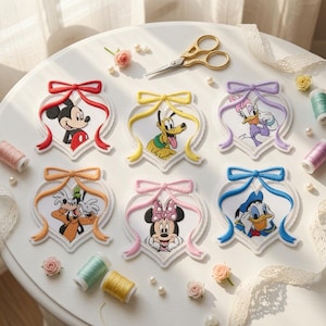 May include: Six embroidered patches shaped like hearts with bows, featuring Mickey Mouse, Pluto, Daisy Duck, Goofy, Minnie Mouse, and Donald Duck. The patches are arranged on a white surface with gold scissors, spools of thread, and small pearls.