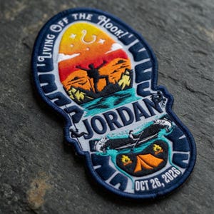 May include: Embroidered patch with a navy blue border and a central design featuring a sunset scene with a person, mountains, and water. The patch includes the text "I'm Living Off The Hook!" and the name "Jordan" with the date "OCT 26, 2026".