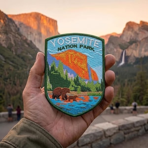 May include: A shield-shaped embroidered patch featuring a landscape scene with a large orange mountain, green pine trees, and two brown bears walking near a blue river. The text YOSEMITE NATION PARK is stitched in white at the top of the patch.