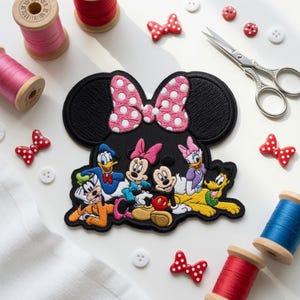 May include: A black embroidered patch shaped like Minnie Mouse ears featuring a pink and white polka dot bow. The center displays a colorful embroidered scene of Goofy, Donald Duck, Minnie Mouse, Mickey Mouse, Daisy Duck, and Pluto.