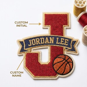 May include: A custom embroidered patch in the shape of the letter "J" with a basketball graphic. The patch is red with a gold border and a navy blue banner that reads "JORDAN LEE". The patch is designed for custom initials and names.
