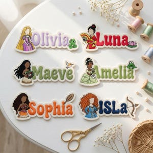 May include: Six embroidered name patches featuring cartoon characters and the names Olivia, Luna, Maeve, Amelia, Sophia, and Isla. Each patch includes a character illustration and a themed icon, such as a frog, lotus, bird, or bow and arrow.