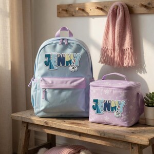 Puede incluir: Una mochila azul claro y una bolsa de almuerzo morado claro a juego, ambas con el nombre JENNY en letras grandes y gr&aacute;ficos de personajes. La mochila tiene un bolsillo frontal, mientras que la bolsa de almuerzo muestra un patr&oacute;n de copos.