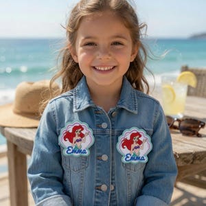 May include: A light blue denim jacket featuring two identical shell-shaped patches on the chest. Each patch displays a cartoon mermaid with long red hair and the name Emma written in blue script lettering below the character on a light background.