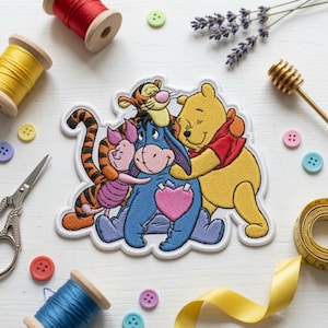 May include: An embroidered fabric patch featuring Winnie the Pooh, Eeyore, Piglet, and Tigger in a group hug. The characters are rendered in vibrant colors with a pink heart on Eeyore's chest. The patch has a white border and is surrounded by sewing supplies.